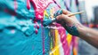 © svastix - An artist's hand, covered in paint, adds layers of bright blue, pink, and yellow to an abstract mural on an outdoor wall, showcasing their vibrant and detailed artistic expression.