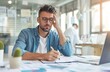 © Cetin - office worker sitting at his desk, looking distressed and holding his head with crumpled paper around him on the table