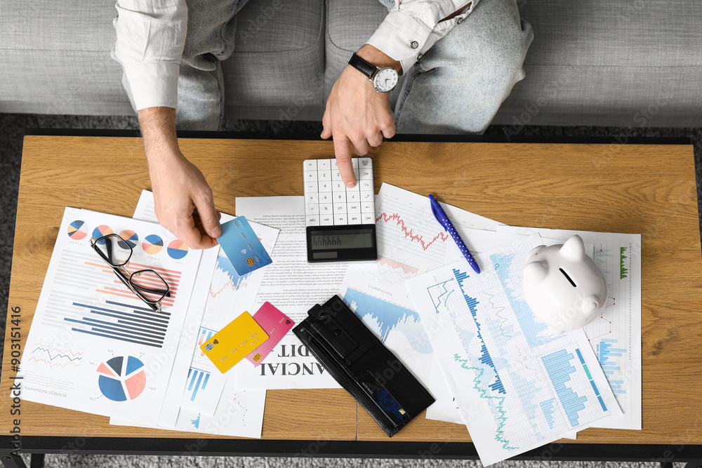 Young man in debt with credit card using calculator at home, top view