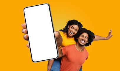 Fototapete - A young black woman in a yellow shirt is giving a piggyback ride to a man in a red shirt, both smiling broadly. The woman is pointing at the blank screen of a large smartphone