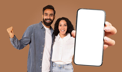 Fototapete - A happy multiracial couple is standing in front of a brown background, holding a large smartphone with a blank screen.