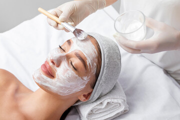 beautician applying white clay mask to face of beautiful young woman in beauty salon. spa treatments