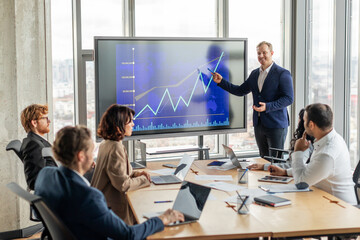 Aufkleber - A businessman stands in front of a large screen displaying a blue line graph that is trending upward, pointing to screen with a pen as he explains positive results to a group of attentive colleagues