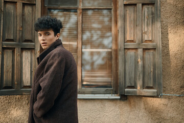 Wall Mural - Fashionable young man in brown coat standing in front of old building with shuttered windows and wooden shutters in background