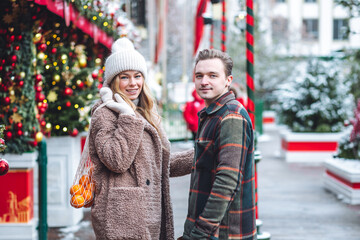 Wall Mural - Beautiful young loving couple embracing, kissing, enjoying winter vacation, cold snowy day on a street city Christmas market outdoors. Warm clothes, knitted sweater, mittens. Cozy atmosphere