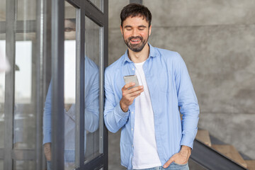 Aufkleber - A smiling man in a light blue shirt and white t-shirt is standing by a glass door, looking down at his smartphone.