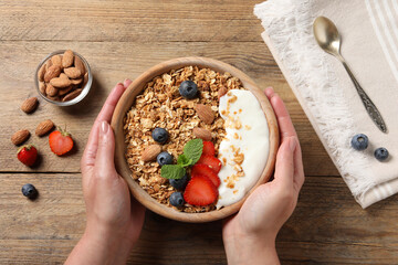 Wall Mural - Woman holding bowl of tasty granola with berries, almond and yogurt at wooden table, top view