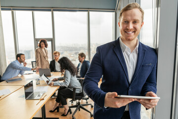 Poster - A smiling businessman stands confidently in a modern office setting, holding a tablet while other colleagues work at a nearby table. The office is brightly lit with windows overlooking a city skyline.