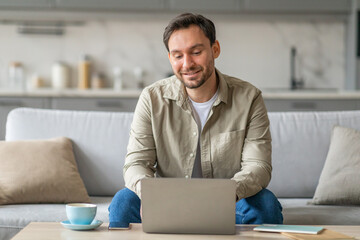 Poster - A man sits on a light gray sofa in a modern living room, working on a laptop computer. He is smiling and focused on his work. There is a cup of coffee on the coffee table in front of him.