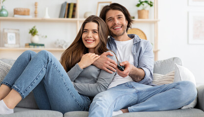 Wall Mural - Joyful young couple watching TV together at home, embracing and sitting on couch. Cheerful man and woman switching channels on TV with remote, seeking nice movie for weekend, empty space