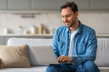 Wall Mural - Man in a blue button-down shirt sits on a white couch in a living room. He is smiling and working on a laptop, which is resting on his lap. He appears to be focused on his work and enjoying his time.