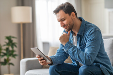 A man wearing a blue denim shirt sits on a gray couch in a living room. He is using a tablet computer and has a thoughtful expression on his face. There is a large lamp in the background.