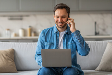 Poster - A man is sitting on a white couch in a living room. He is wearing a blue denim shirt and blue jeans. He is smiling as he talks on a phone held to his ear with his right hand, looking at laptop