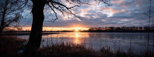  Sun sets over water with trees in foreground, cloudy sky in background