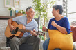 © ztony1971 - An elderly man plays the guitar while his wife listens with a smile, sharing a tender moment of music and companionship in their cozy living room.