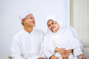 Wall Mural - Muslim little girl chatting with her grandparents while sitting together on the couch during Eid Mubarak in living room at home