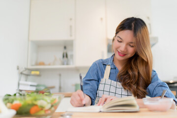 Wall Mural - Young asian woman writing food recipes in notebook and eating yogurt while preparing fresh vegetables salad and cooking healthy breakfast food in modern kitchen with healthy lifestyle at home