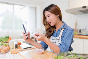 Wall Mural - Young asian woman eating yogurt and use smartphone to searching food recipes while preparing fresh vegetables salad and cooking healthy breakfast food in modern kitchen with healthy lifestyle at home