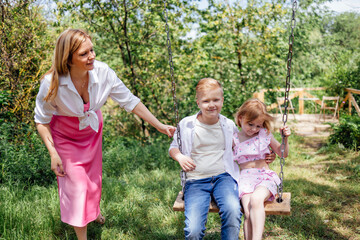 Young attractive mom rides her cute children on a swing in a meadow in the park.