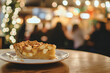 © Anastasia - Freshly baked apple pie slice on a white plate sits on a wooden table, with a blurred background of warm lights and people enjoying a cozy atmosphere