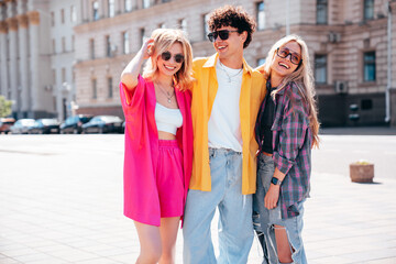 Wall Mural - Group of young three stylish friends posing in the street. Fashion man and two cute female dressed in casual summer clothes. Smiling models having fun. Cheerful women and guy outdoors, in sunglasses