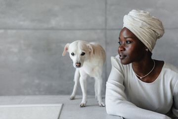 contemplative woman in a white outfit and head wrap sits beside a small white dog, both gazing thoughtfully in a modern interior, portraying a serene and peaceful moment