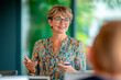 © GustavsMD - A senior businesswoman with glasses and short hair leads a meeting with enthusiasm, engaging with participants in a modern office.