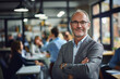 © GustavsMD - A confident senior professional man with glasses and a beard stands with arms crossed in a busy, modern office environment with colleagues working in the background.