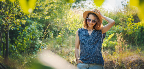 Wall Mural - Summer portrait of cheerful young woman in hat and sunglasses walking in the park