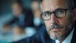 ©  lukaPixMedia - A serious man with gray hair and glasses, dressed in formal attire, gazes intensely, with blurred colleagues and corporate setting in the background.