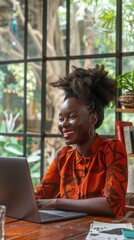 Wall Mural - Vibrant workspace scene: woman in bright orange shirt smiles while working on laptop by large window, lush greenery visible outside, natural light illuminates cozy interior.