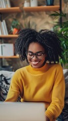 Wall Mural - Radiant woman with natural curly hair and glasses beams while working on laptop in cozy home office, her yellow sweater adding warmth to the studious atmosphere.