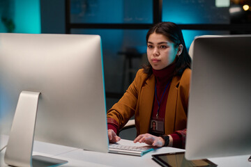 Portrait of successful young woman as female cybersecurity expert using computer in office and looking at screen with blue light
