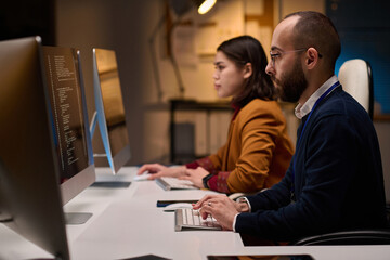 Side view portrait of two software developers using computers and writing code sitting at workplace in office