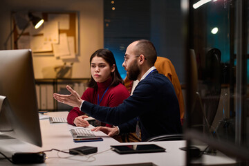 Side view portrait of bearded software developer advising young colleague and pointing at computer screen while collaborating in office