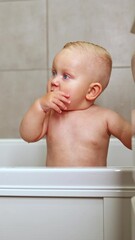 Wall Mural - A cute blue-eyed 1-year-old boy is bathing in a white bath. Baby's personal hygiene. A child with a smile on his face while bathing. Vertical video