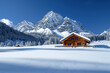 © LarisaM - Charming wooden cabin nestled in a snowy mountain landscape, surrounded by pine trees and under a clear blue sky.