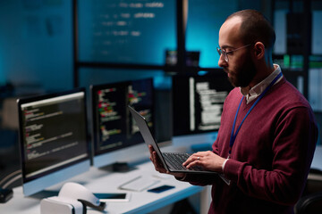 Side view portrait of bearded adult man as computer programmer holding open laptop in IT and cybersecurity office with blue lights copy space