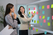 © PaeGAG - Businesswomen Collaborating in Modern Office with Colorful Sticky Notes on Whiteboard