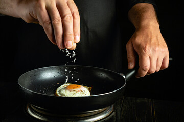 Wall Mural - The chef fries an egg in a frying pan. Adding salt to the eggs by the chef hands. Healthy breakfast from scrambled eggs.