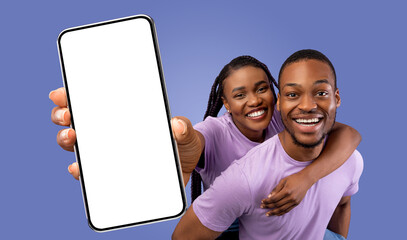 Fototapete - A happy black couple poses for a picture while holding up a smartphone with a blank screen. They are both smiling and looking at the camera.
