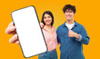 © Prostock-studio - A young man and woman stand in front of a yellow background. The woman is holding a smartphone with a blank white screen towards the viewer, while the man gives a thumbs up. Both are smiling.