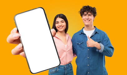 Fototapete - A young man and woman stand in front of a yellow background. The woman is holding a smartphone with a blank white screen towards the viewer, while the man gives a thumbs up. Both are smiling.
