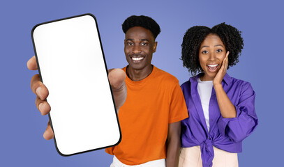 Fototapete - African American man and woman are standing side by side in front of a blue background. The man is holding out a smartphone with a blank screen towards the viewer