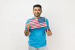 © khosrork - Portrait of cheerful unshaven man wearing blue T- shirt standing holding flag of United States of America, looking at camera with toothy smile. Indoor studio shot isolated on gray background.