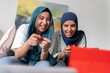 © Studio Marmellata - Two women in hijabs playing video games with excitement on a handheld console, showing expressions of joy and surprise, creating a lively and interactive environment