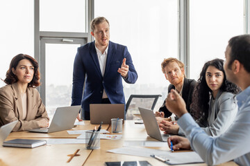 A group of business professionals are gathered around a conference table in a modern office. The presenter, standing at the head of the table, is speaking with passion and enthusiasm