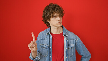 Young man with curly hair wearing a denim jacket and red shirt posing against a red background, index finger raised