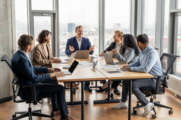 Aufkleber - A group of five professionals are seated around a large, rectangular table in a modern office. They are engaged in a discussion, looking at each other and using laptops