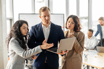 Aufkleber - Three business professionals stand in a modern office, gathered around a laptop. They are all attentively listening and appear to be engaged in a productive conversation.